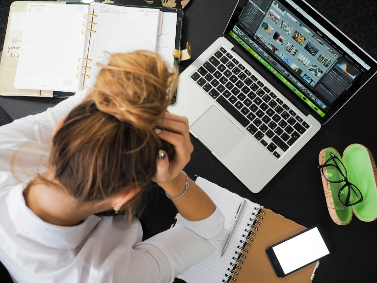 Eine Frau sitzt an einem Schreibtisch mit offenem Laptop und stützt ihren Kopf auf den Händen ab. Um sie herum liegen ein Notizheft und ein Terminkalender.