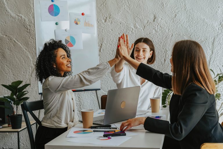 Drei fröhliche Business-Frauen sitzen an einem Tisch und feiern ihre Erfolge, indem sie die Hände abklatschen.